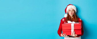 快乐假期圣诞节概念可爱的红色头发的人女孩持有礼物皱<strong>纹嘴唇</strong>吻穿圣诞老人他有趣的毛衣蓝色的背景
