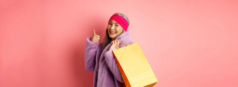 时尚的亚洲高级女人购物携带纸袋肩膀显示翘拇指推荐商店折扣粉红色的背景