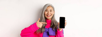 在线购物美丽的亚洲祖母微笑指出手指智能手机空白屏幕显示移动应用程序站白色背景
