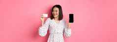快乐年轻的亚洲女人互联网购物显示智能手机屏幕满意信贷卡站粉红色的背景