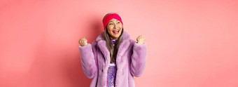 亚洲高级女人庆祝欢呼赢得使拳头泵迹象胜利实现目标微笑满意站时尚的人造皮毛外套粉红色的背景