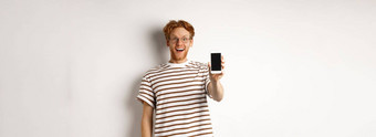 技术电子商务概念快乐年轻的红色头发的人男人。眼镜显示空白智能<strong>手机屏</strong>幕相机惊讶站白色背景