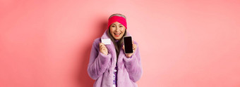 在线购物时尚概念美丽的亚洲成熟的女人显示塑料信贷卡空白电话屏幕微笑快乐粉红色的背景