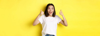 漂亮的年轻的亚洲女人白色t恤显示拇指微笑赞美好提供推荐产品站满意黄色的背景