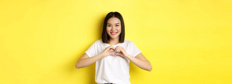美丽的亚洲女人显示<strong>爱心手势</strong>微笑相机站黄色的背景概念情人节一天浪漫