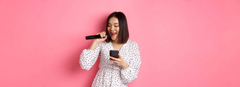 可爱的亚洲女人阅读歌词智能手机唱歌麦克风站时尚的衣服粉红色的背景