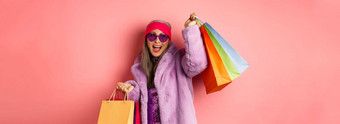 时尚的亚洲高级女人购物穿时尚的衣服太阳镜持有商店袋跳舞快乐粉红色的背景