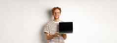 微笑骄傲红色头发的人的家伙显示空移动PC屏幕英俊的年轻的男人。展示标志电脑显示白色背景