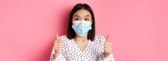 科维德流感大流行生活方式概念支持亚洲女脸面具显示竖起大拇指批准赞美好工作站粉红色的背景
