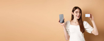 年轻的高加索人女人显示塑料信贷卡很高兴微笑演示空<strong>手机屏</strong>幕站米色背景