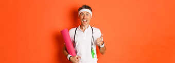 概念锻炼健身房生活方式快乐的中年健身的家伙持有跳过绳子瑜伽席显示竖起大拇指微笑满意站橙色背景