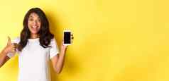 肖像兴奋非裔美国人女孩显示智能手机屏幕使竖起大拇指推荐很酷的站黄色的背景