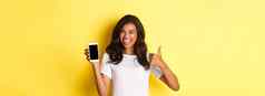 图像美丽的非裔美国人女孩眨眼微笑显示智能手机屏幕竖起大拇指批准站黄色的背景