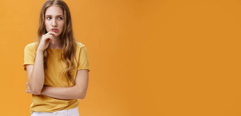 肖像有关聪明的女人波浪公平头发休闲t恤持有手下巴追求嘴唇深思熟虑的令人<strong>担忧</strong>的表达式摆姿势陷入困境的橙色墙