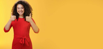 有创意的有魅力的快乐乐观的女人卷曲的头发红色的衣服眨眼批准显示拇指广泛的微笑满意给积极的回复黄色的墙
