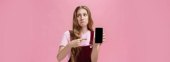 室内拍摄悲观的不高兴失望可爱的年轻的女孩持有智能手机指出手机屏幕使心烦意乱鬼脸困扰不满意粉红色的墙