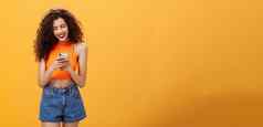 女孩支出时间互联网发短信朋友消息智能手机笑设备屏幕站快乐乐观的橙色背景裁剪前牛仔布短裤