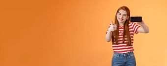 兴奋快乐的红色头发的人有吸引力的女人很高兴显示拇指介绍太棒了手机游戏持有智能手机水平微笑批准推荐好在线商店橙色墙