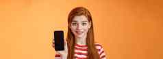 特写镜头可爱的高加索人红色头发的人女孩显示智能手机屏幕橙色背景微笑可爱的推荐应用程序促进应用程序在线提供出售介绍移动电话
