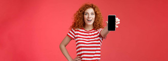 快乐的有吸引力的有魅力的欧洲红色头发的人女孩卷曲的发型显示智能手机屏幕微笑幸福的促进应用程序建议推荐好应用程序社会媒体页面红色的背景