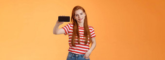 时髦的自信有吸引力的高加索人姜女孩介绍智能手机应用程序推荐很酷的游戏显示手机屏幕水平微笑骄傲吹牛<strong>打分</strong>数站橙色背景
