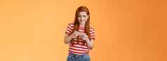 兴奋红色头发的人高加索人女孩强烈的皱着眉头通过硬游戏水平玩商场利用屏幕持有智能手机水平电话显示谨慎的橙色背景