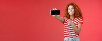 兴奋快乐好看的红色头发的人卷曲的女显示水平智能手机屏幕指出显示小工具微笑很高兴骄傲击败朋友分数游戏站红色的背景
