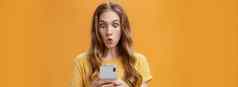 年轻的女孩震惊了阅读消息朋友智能手机折叠嘴唇出现眼睛惊讶的是手机屏幕折叠嘴唇感兴趣兴奋摆姿势橙色背景