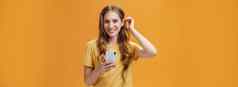 半身照拍摄可爱的友善的不错的年轻的女孩波浪发型把头发海滩耳朵持有智能手机微笑广泛的快乐的表达式相机橙色背景