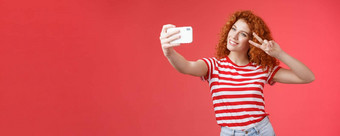 时尚的无忧无虑的好看的红色头发的人卷曲的女人享受夏天假期显示胜利和平手势眼睛倾斜头可爱的记录视频持有智能手机采取自拍红色的背景