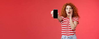 逗乐深刻的印象红色头发的人好看的卷发兴奋女孩分享太棒了社会媒体页面显示智能手机屏幕惊讶触摸脸颊激动影响太棒了应用程序红色的背景