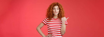 的东西红色头发的人好奇的时髦的好看的卷发女孩夏天条纹t恤傻笑盯着指出左指示拇指有趣的产品站红色的背景