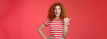 时髦的好看的感情的快乐微笑红色头发的人欧洲女卷曲的发型指出拇指左露齿而笑自信的厚颜无耻的持有手腰导演<strong>促销广告</strong>提供红色的背景