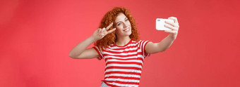 时尚的无忧无虑的好看的红色头发的人卷曲的女人享受夏天假期显示胜利和平手势眼睛倾斜头可爱的记录视频持有智能<strong>手机</strong>采取自拍红色的背景