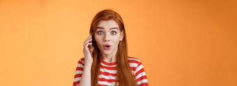 逗乐激动红色头发的人女孩发现难以置信的新闻会说话的电话持有智能手机耳朵站在相机惊讶惊讶告诉太棒了促销站惊讶橙色背景