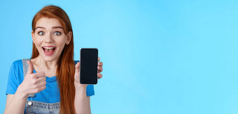 惊讶着迷可爱的红色头发的人女人推荐好智能<strong>手机</strong>太棒了应用程序电话游戏<strong>显示手机显示</strong>激动相机微笑批准很酷的设备蓝色的背景
