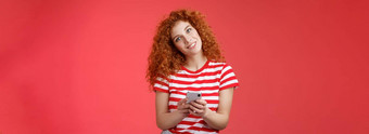 愚蠢的可爱的红色头发的人卷发时尚女持有智能手机倾斜头微笑广泛的露出牙齿的积极的露齿而笑思考回答发送挑衅时髦的消息红色的背景