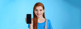 特写镜头有吸引力的快乐的红色头发的人年轻的女商店助手建议客户买智能手机显示电话显示促进应用程序互联网应用程序微笑相机站蓝色的背景