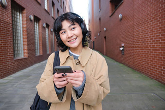 肖像年轻的朝鲜文女人走城市背包耳机听音乐智能手机电话应用程序街道