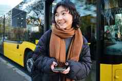 年轻的美丽的女人站公共汽车停止发短信消息智能手机持有移动电话检查时间表购买票在线穿冬天衣服