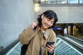 肖像可爱的朝鲜文女孩沟外套自动扶梯持有移动<strong>电话</strong>微笑很高兴通勤手表视频听音乐无线耳机