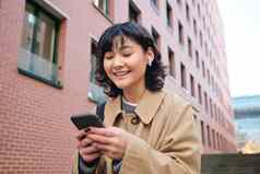 快乐朝鲜文女孩走街听音乐无线耳机持有智能手机选择首歌播放列表站户外建筑阅读消息