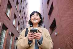 较低的角视图浅黑肤色的女人朝鲜文女孩听音乐耳机走街智能手机阅读消息移动电话