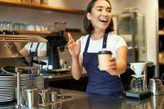 肖像微笑亚洲女孩咖啡师给订单咖啡馆邀请客人选择外卖订单计数器持有外卖杯咖啡