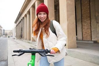微笑红色头发的人欧洲女孩驱动器公共e-scooter旅游探讨了城市游乐设施城市中心
