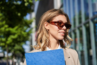 关闭肖像微笑年轻的女人太阳镜企业西装持有文件夹工作文档面试站在户外