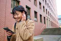 微笑浅黑肤色的女人女人亚洲学生走街听音乐无线耳机持有智能手机读取文本消息移动电话选择首歌播放列表