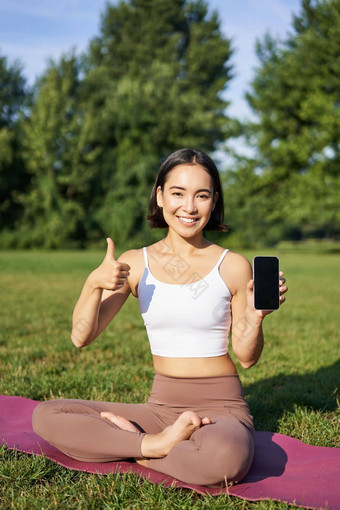 垂直拍摄亚洲女人显示拇指<strong>推</strong>荐瑜伽<strong>培训</strong>在线冥想应用程序练习新鲜的空气公园显示智能手机屏幕