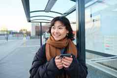 肖像朝鲜文女人冬天夹克站智能手机等待公共汽车停止移动电话应用程序检查公共运输应用程序