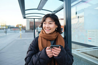 肖像朝鲜文女人冬天夹克站智能<strong>手机</strong>等待公共汽车停止移动电话<strong>应用</strong>程序检查公共运输<strong>应用</strong>程序